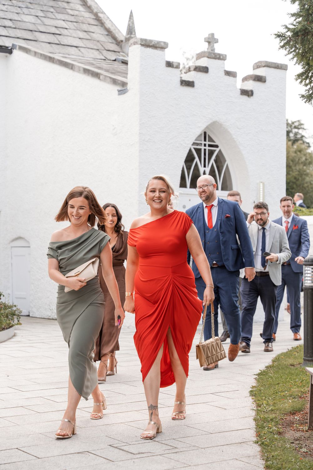 Wedding guests in elegant attire walking from a white church, women in red and olive dresses leading the procession.
