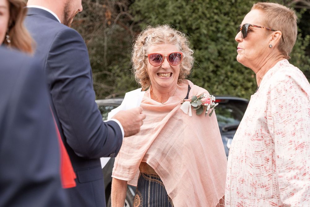 Elderly woman with curly hair and pink sunglasses smiling at an outdoor wedding event with guests nearby.