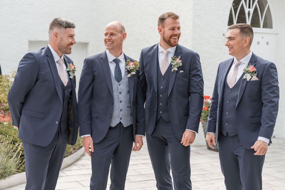 Four groomsmen in navy suits with floral boutonnieres smiling outdoors at a wedding celebration.