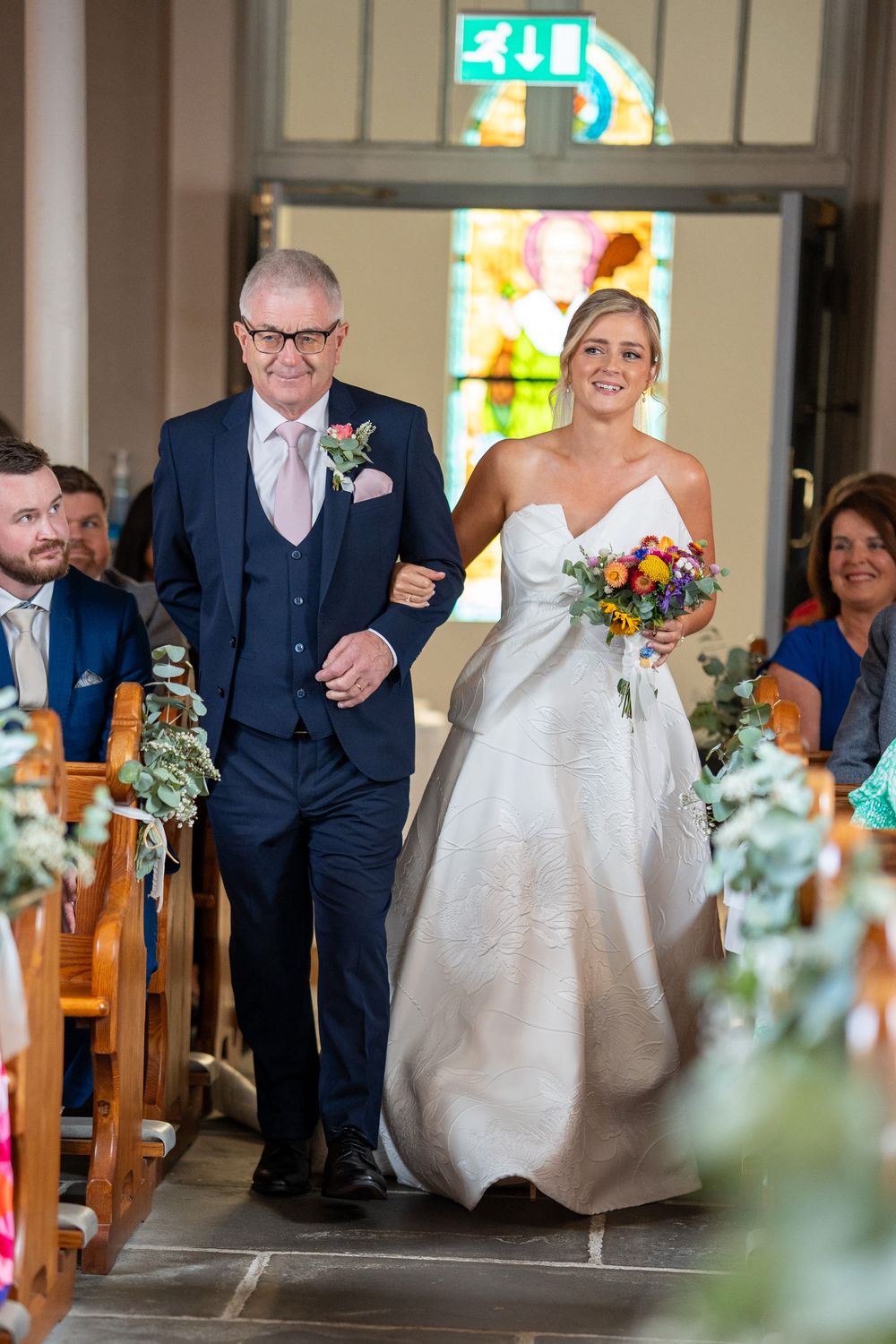 Bride in white gown walks down church aisle with older man in navy suit, holding colorful bouquet.