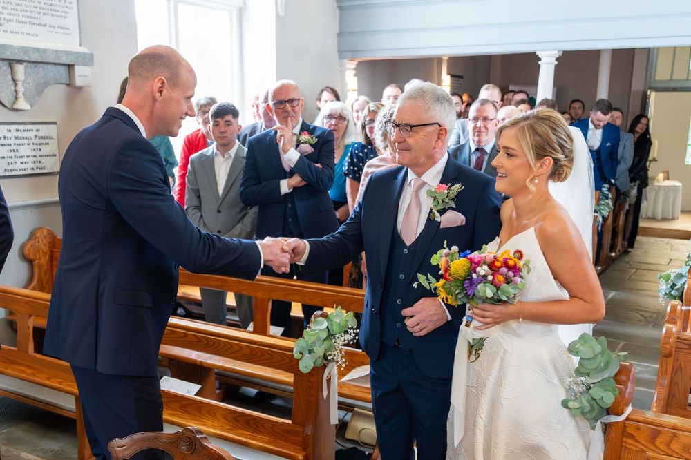 Bride and groom shaking hands with guest at church wedding ceremony aisle, surrounded by seated congregation.