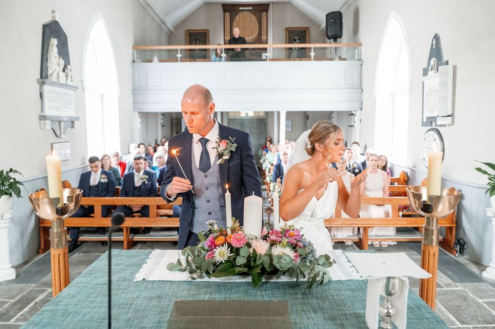 Bride and groom lighting unity candles at church altar during wedding ceremony with guests seated in pews behind them.