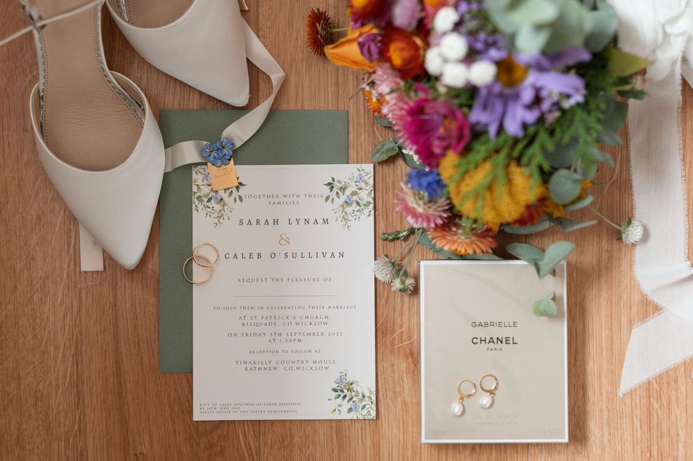 Elegant wedding flat lay with sage green invitation suite, gold rings, Chanel earrings, bridal heels, and colorful floral bouquet.