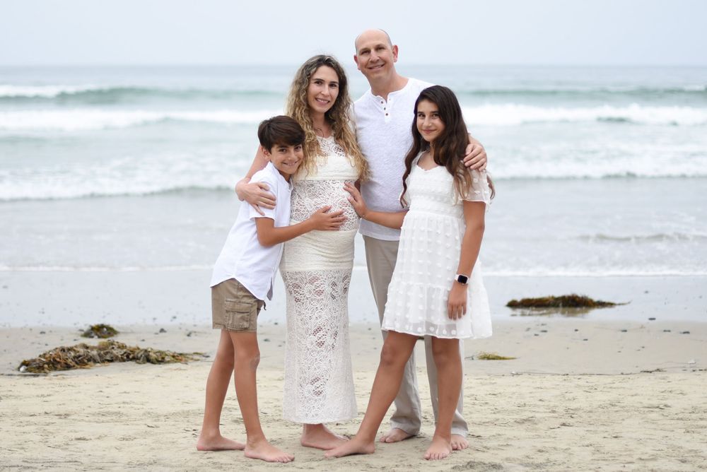 Family of four posing together on a sandy beach, wearing white outfits, with ocean waves in the background.