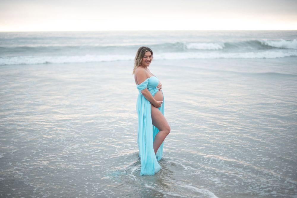 Pregnant woman in teal maternity gown standing in ocean waves at beach during golden hour photography session.