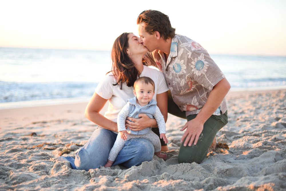 Happy family with baby sitting on sandy beach, parents kissing while holding infant, ocean waves in background.
