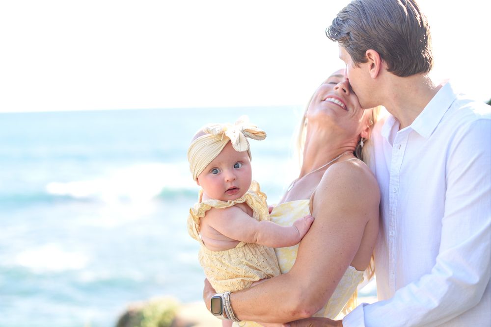 A young family with a baby girl in a yellow outfit poses on a rocky beach with ocean waves in the background.