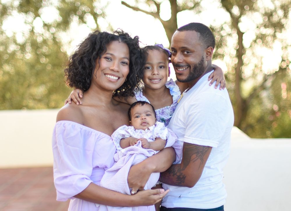 Happy family of four posing outdoors, parents holding newborn baby and young daughter, smiling together.