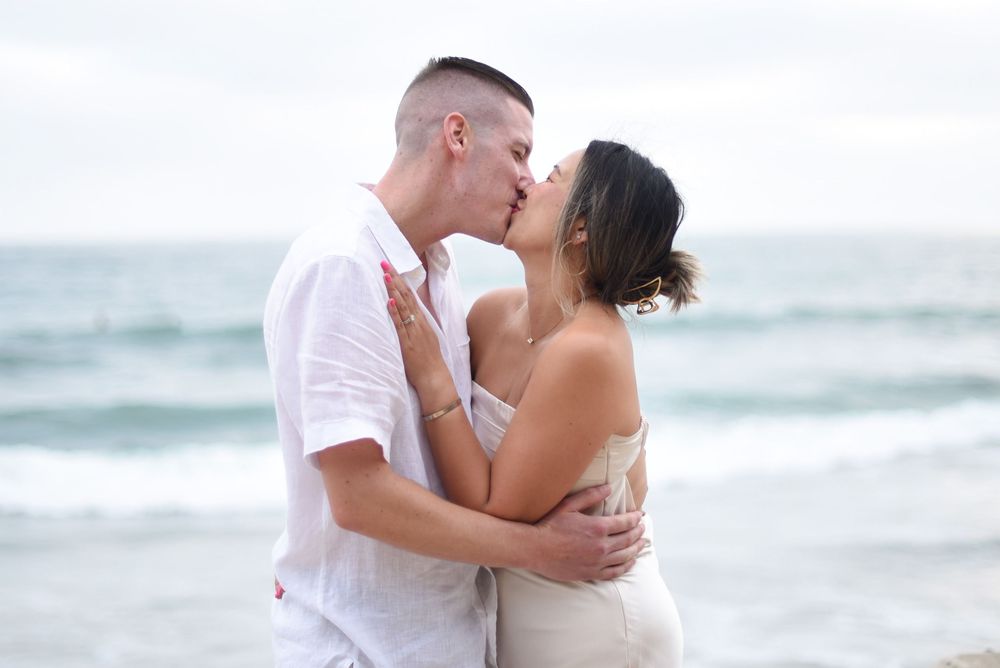 Couple sharing a romantic kiss on the beach, dressed in white, with ocean waves in the background.