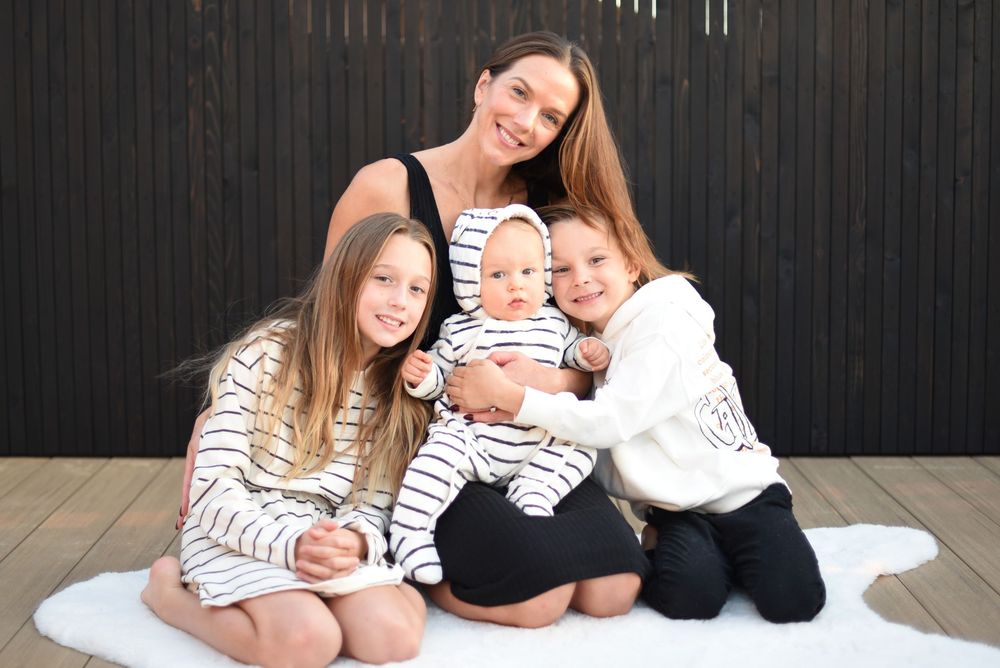 A smiling mother poses with three young children in matching black and white striped outfits on a white rug outdoors.