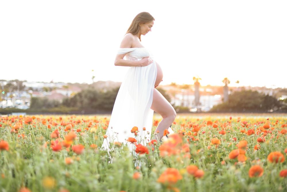Pregnant woman in white dress standing in vibrant orange poppy field during golden hour sunset.