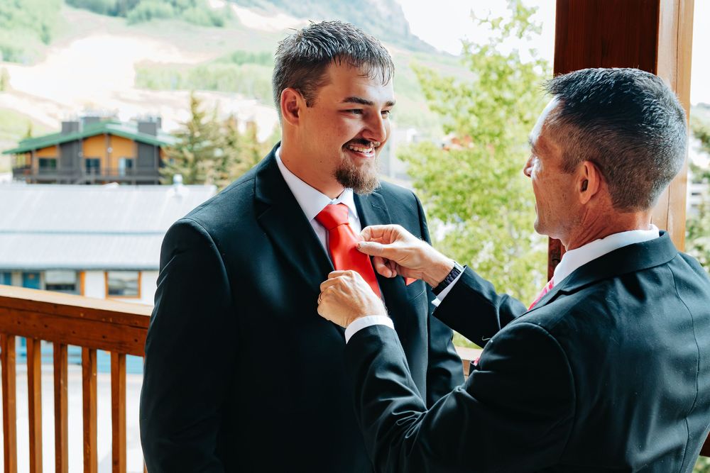 Smiling groom having red tie adjusted by man on outdoor balcony with mountain views.