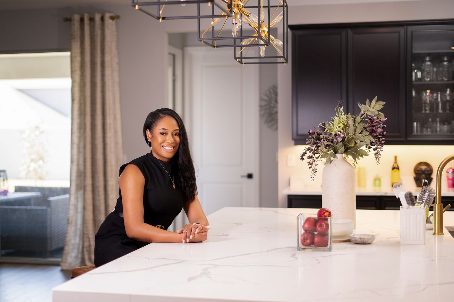 A smiling woman in a black top leans on a white marble kitchen counter with gold pendant lights above.