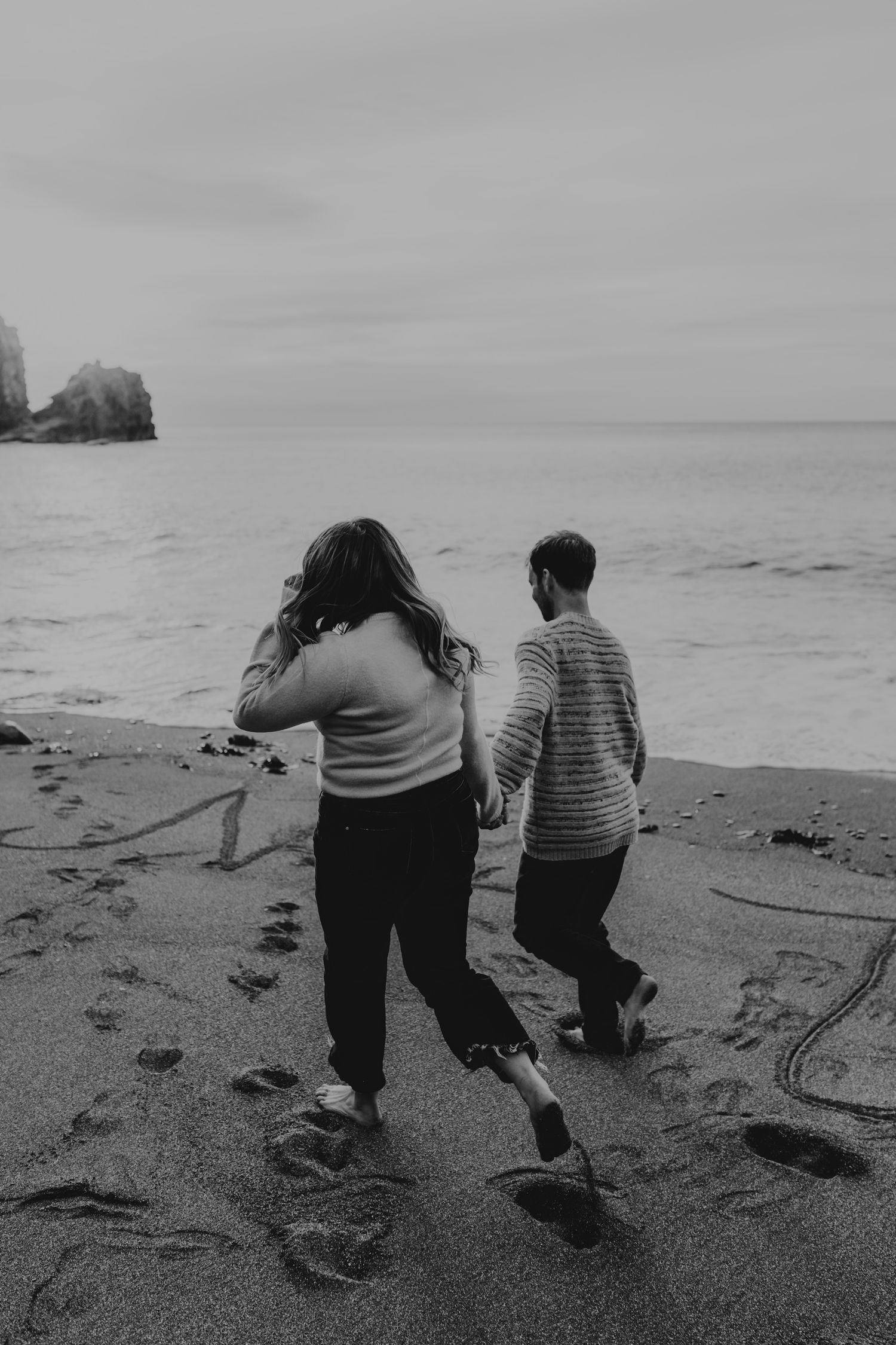 Black and white photo of a couple holding hands walking on a sandy beach toward ocean waves, with footprints in the sand.