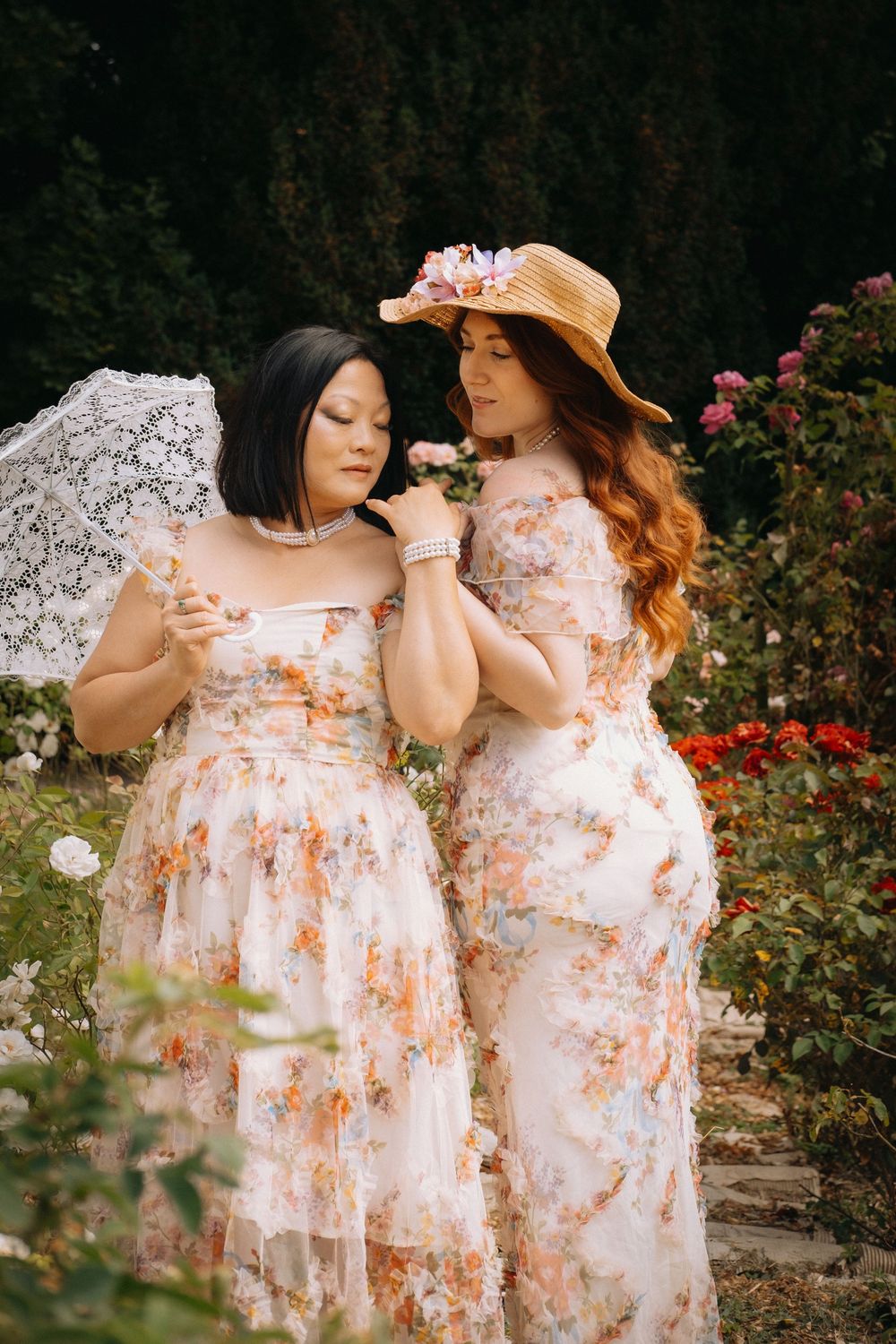 Portrait de deux femmes en robes fleuries dans un jardin, entourées de végétation et de fleurs.