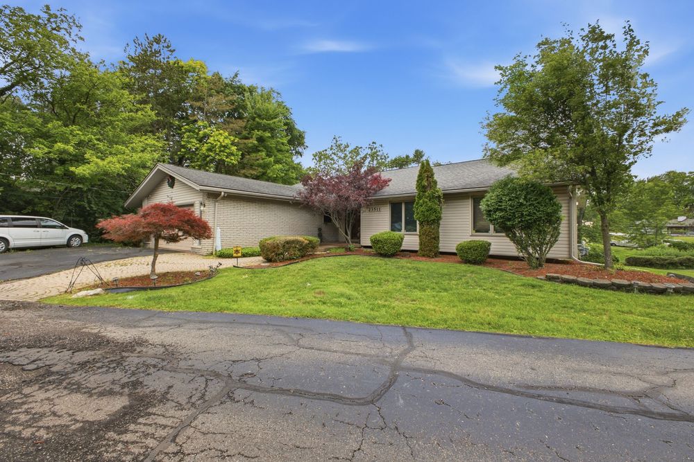 Ranch-style home with lush green lawn, mature trees, red-leaf Japanese maple, and paved driveway on a sunny day.