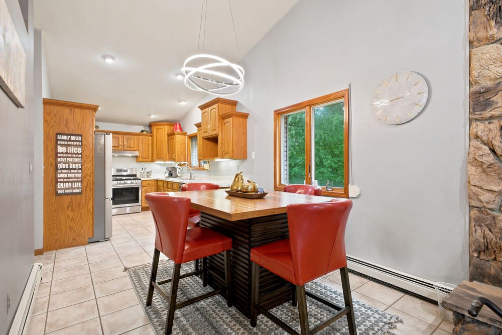 Bright kitchen and dining area with red chairs, wood table, chandelier, oak cabinets, and a window view of lush greenery.