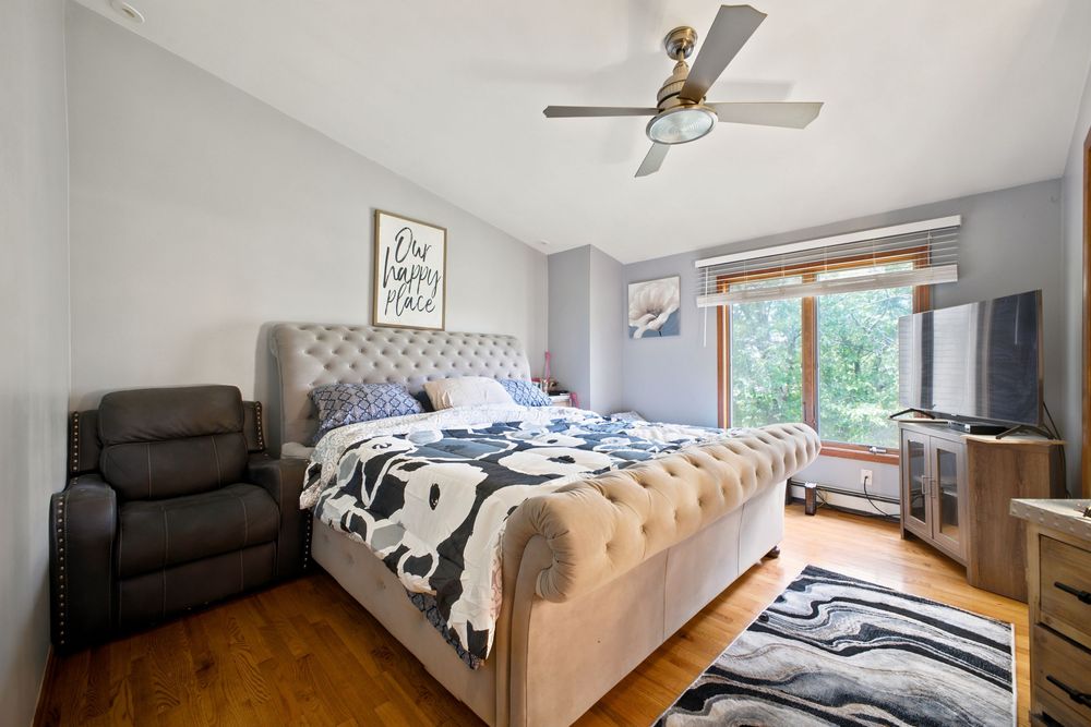 Cozy bedroom with tufted beige bed, hardwood floors, ceiling fan, recliner chair, and large window with garden view.