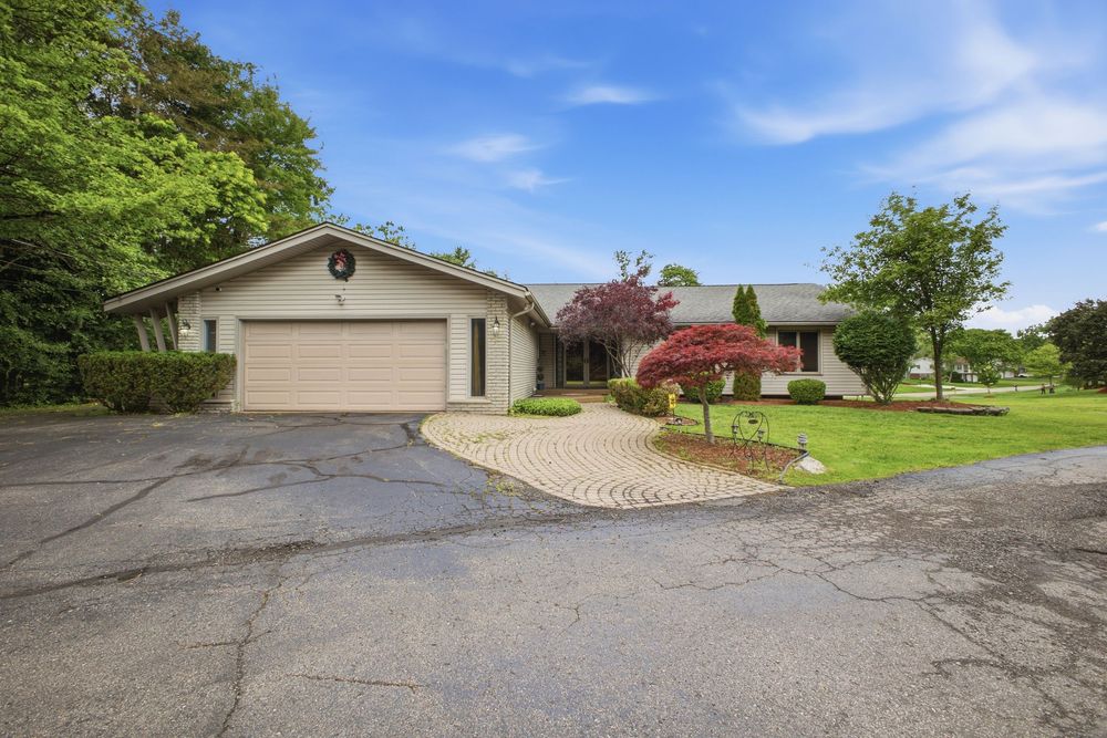 Ranch-style home with attached garage, circular paver driveway, red Japanese maple tree, and lush green lawn surrounded by mature trees.