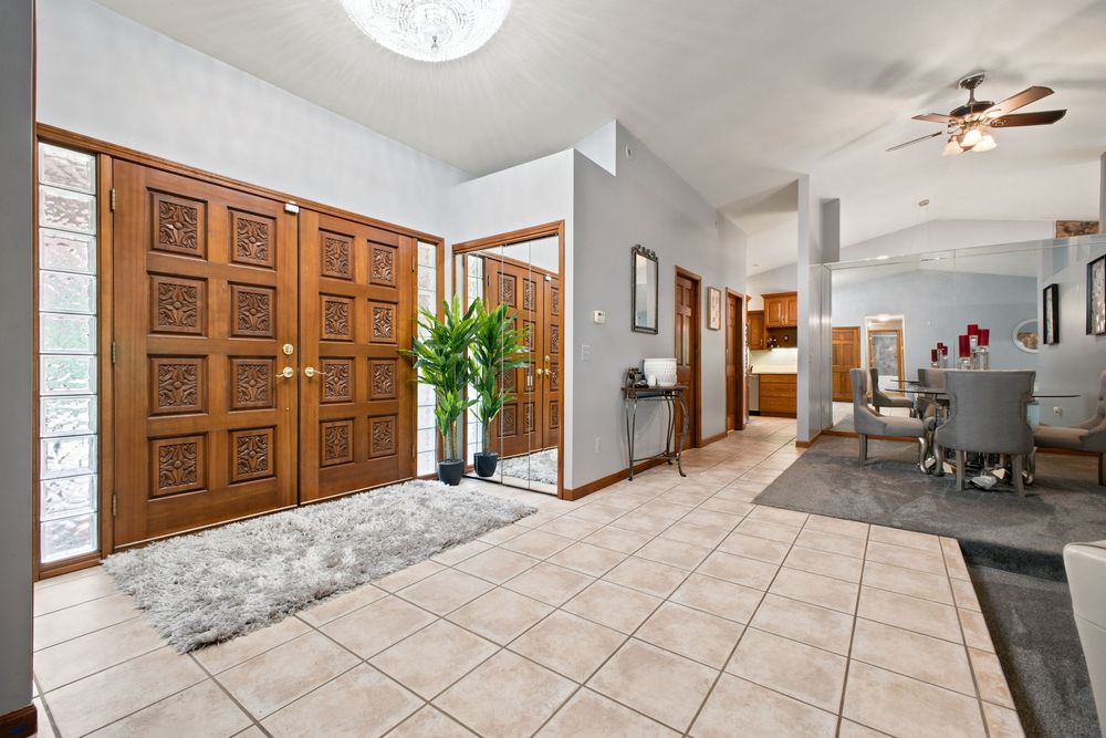 Spacious entryway with ornate wooden double doors, tile flooring, high ceilings, and open view into dining area with ceiling fan.