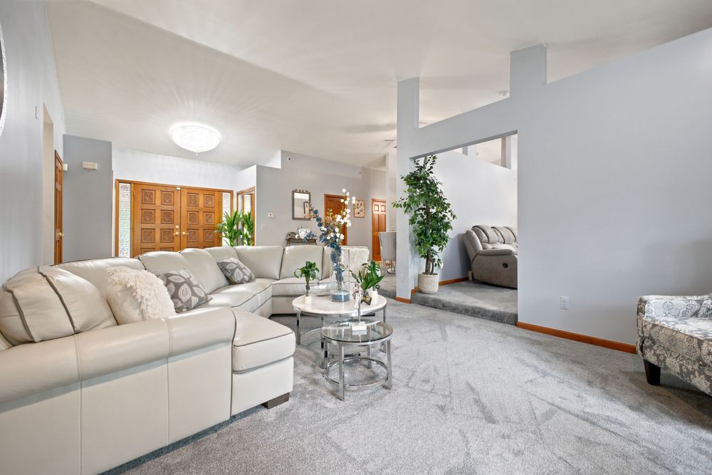 Bright living room with light gray carpet, white sectional sofa, glass coffee table, high ceilings, and wood shuttered windows.