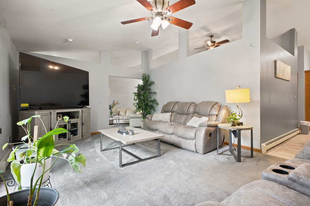 Spacious living room featuring vaulted ceiling, ceiling fan, large TV, sectional sofa, and indoor plants.