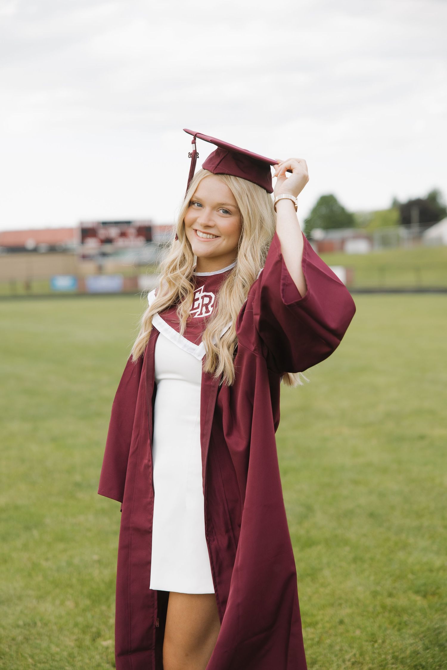 Smiling blonde graduate in maroon cap and gown poses outdoors on green grass field on cloudy day.