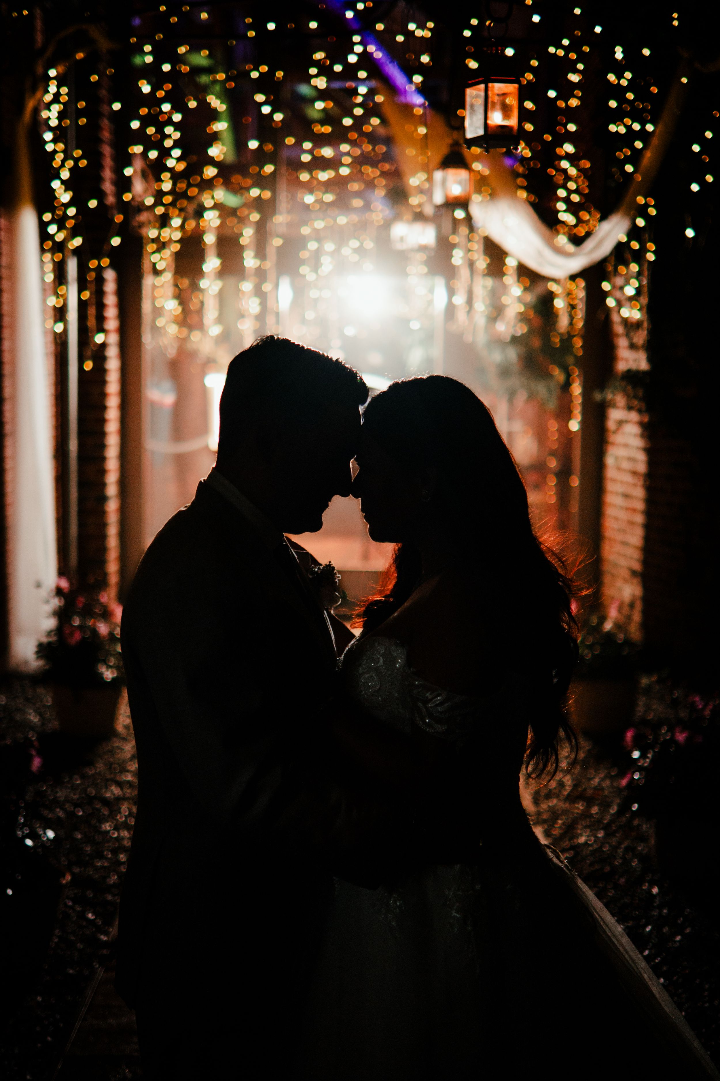 A smiling bride and groom embrace outdoors, with the groom in a beige suit holding the curly-haired bride.