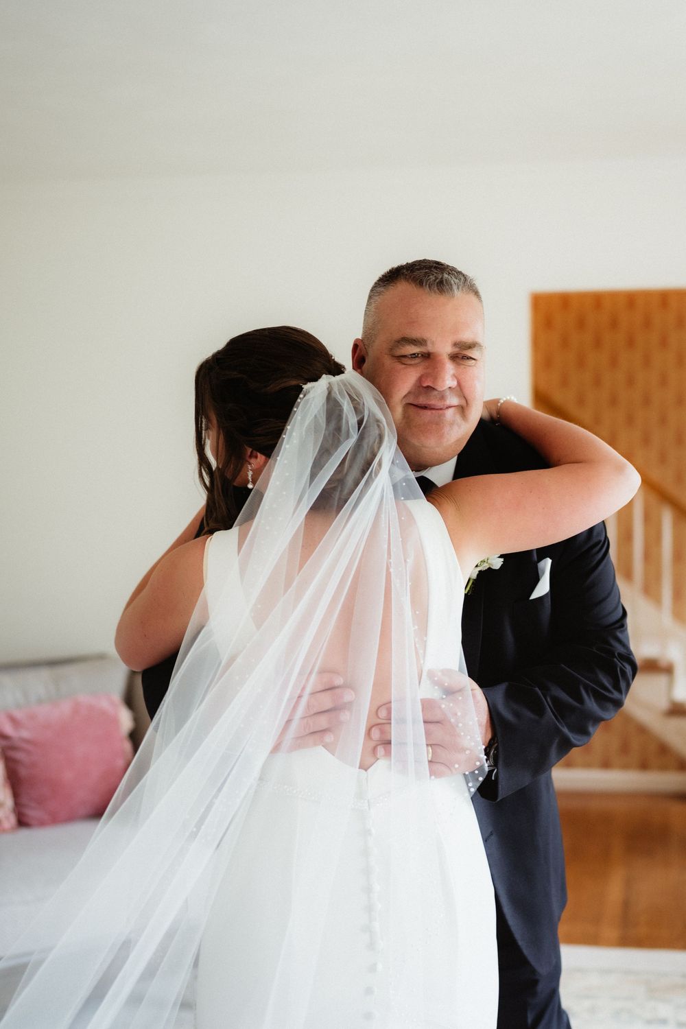 Father embracing bride in wedding dress and veil during emotional first look moment indoors.
