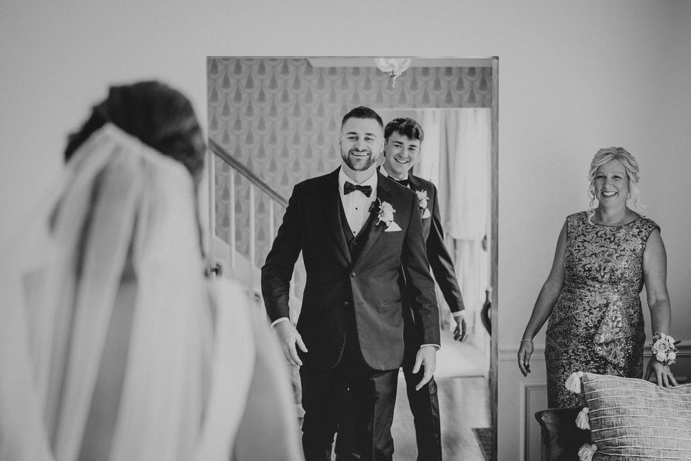 Groom smiling as he sees bride for first time, with guests watching emotional first look moment in black and white.