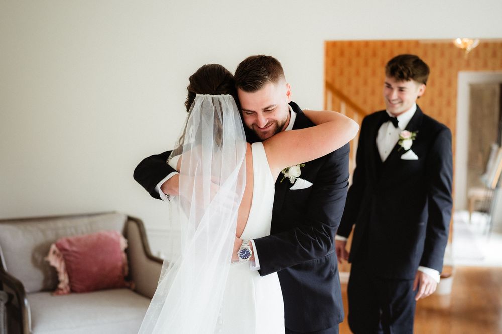Groom embracing bride in wedding dress and veil during emotional first look moment, with groomsman in background.