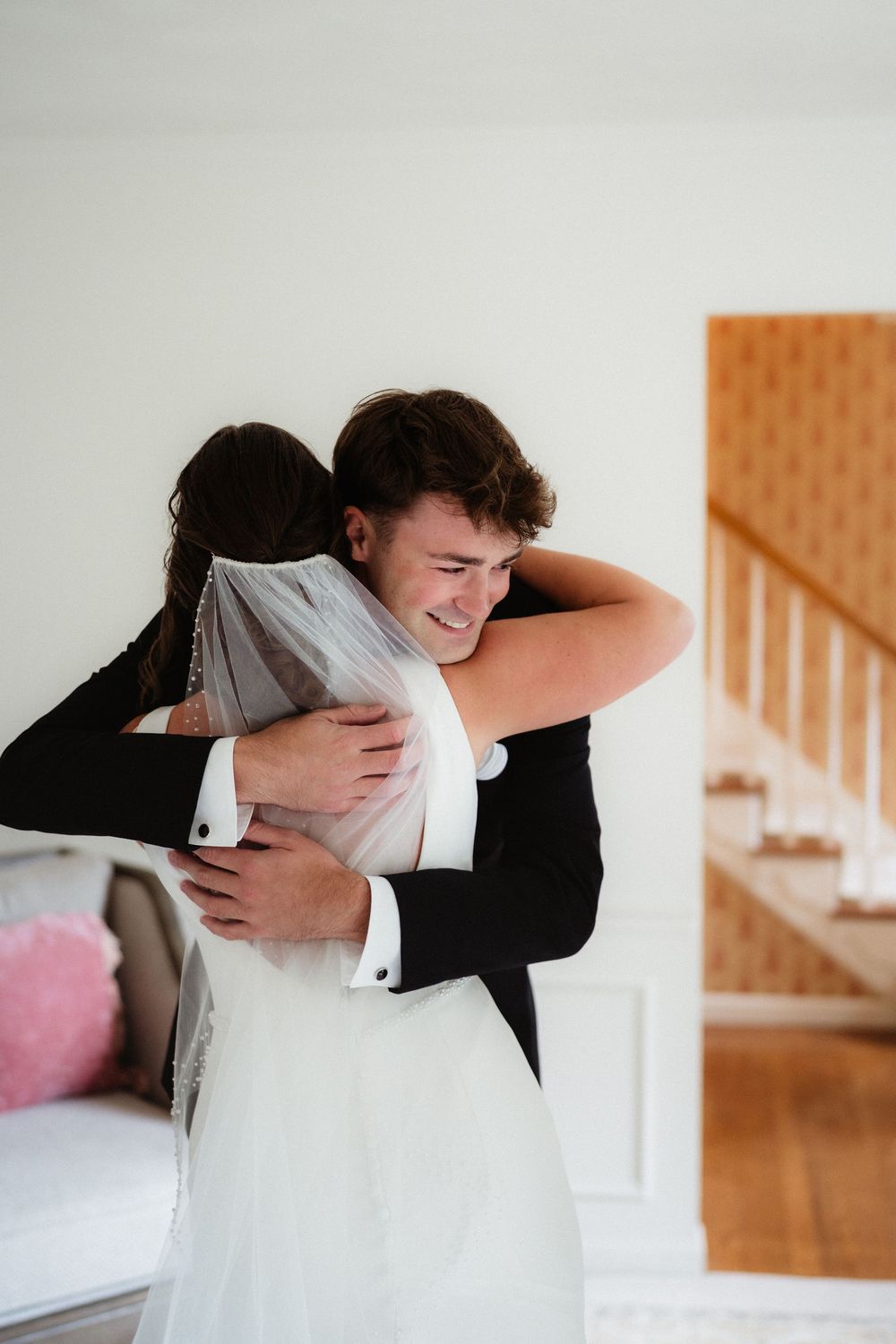 Emotional groom in black tuxedo hugging bride in white wedding dress and veil during first look moment.