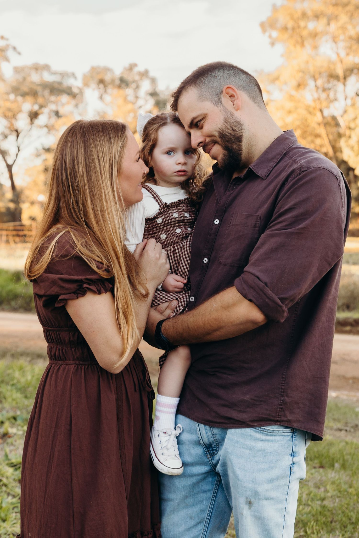 A loving family portrait with parents in brown outfits holding their toddler daughter outdoors at golden hour.