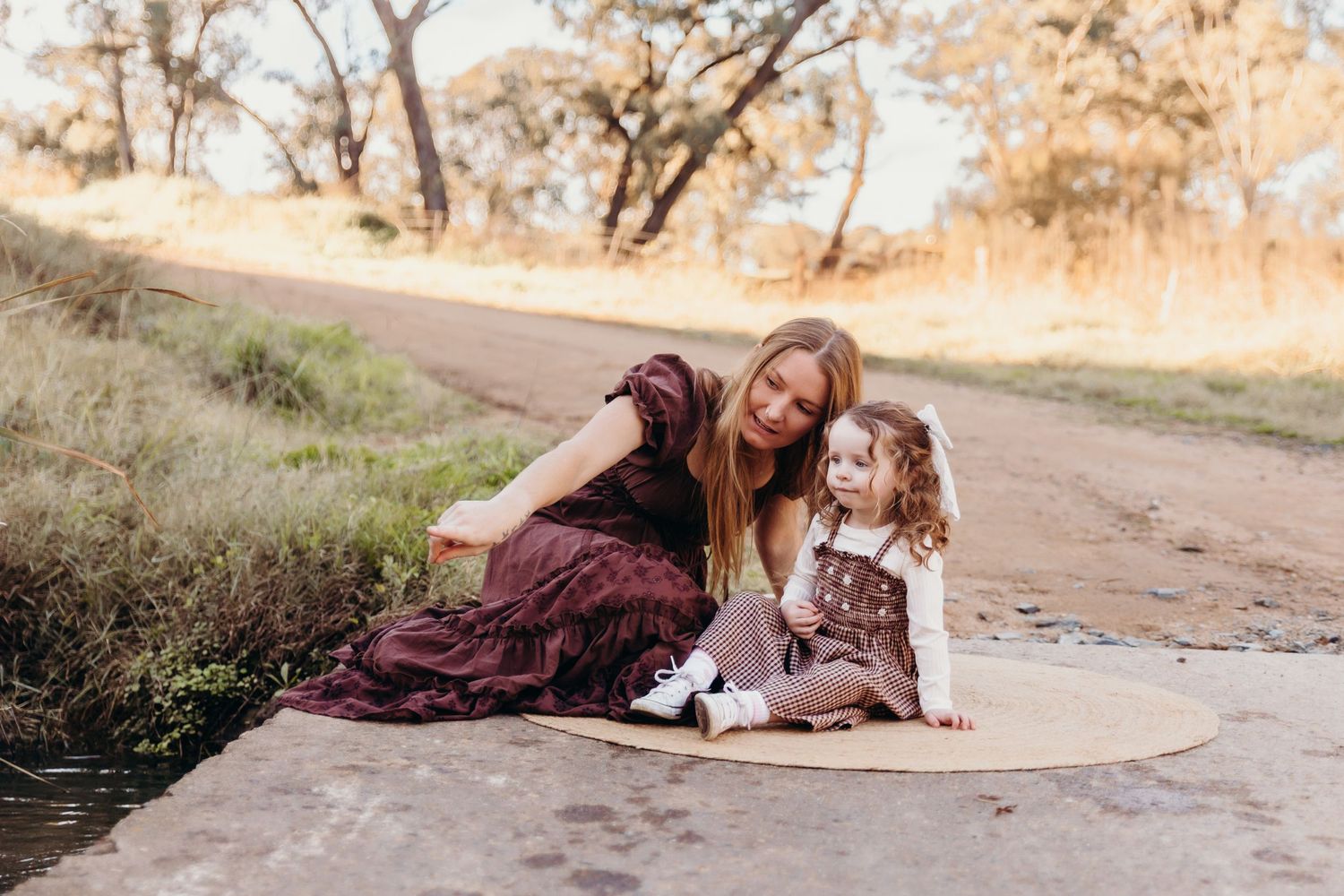 A young mother in a burgundy dress sits with her toddler daughter on a rural dirt path.