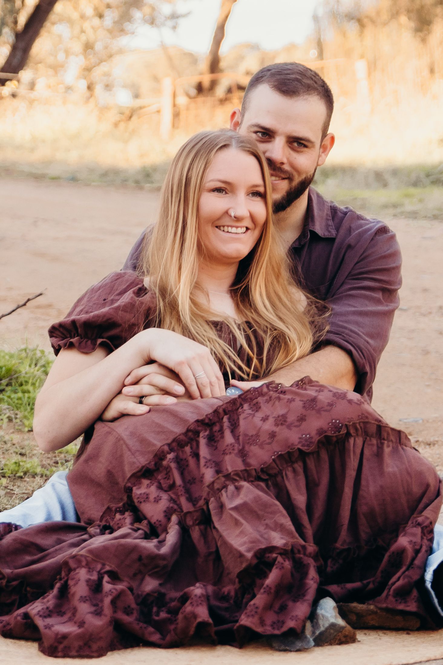 A smiling couple sits together on a dirt path, the man embracing the woman from behind.