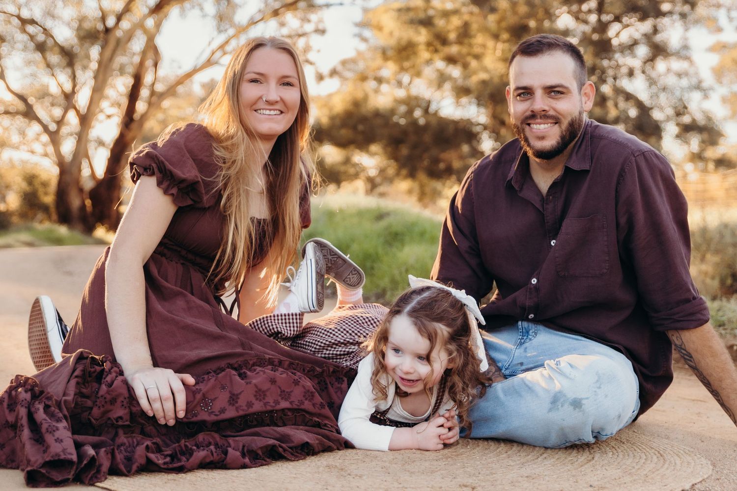 A happy family with a young toddler girl poses together on a sunlit rural dirt road.