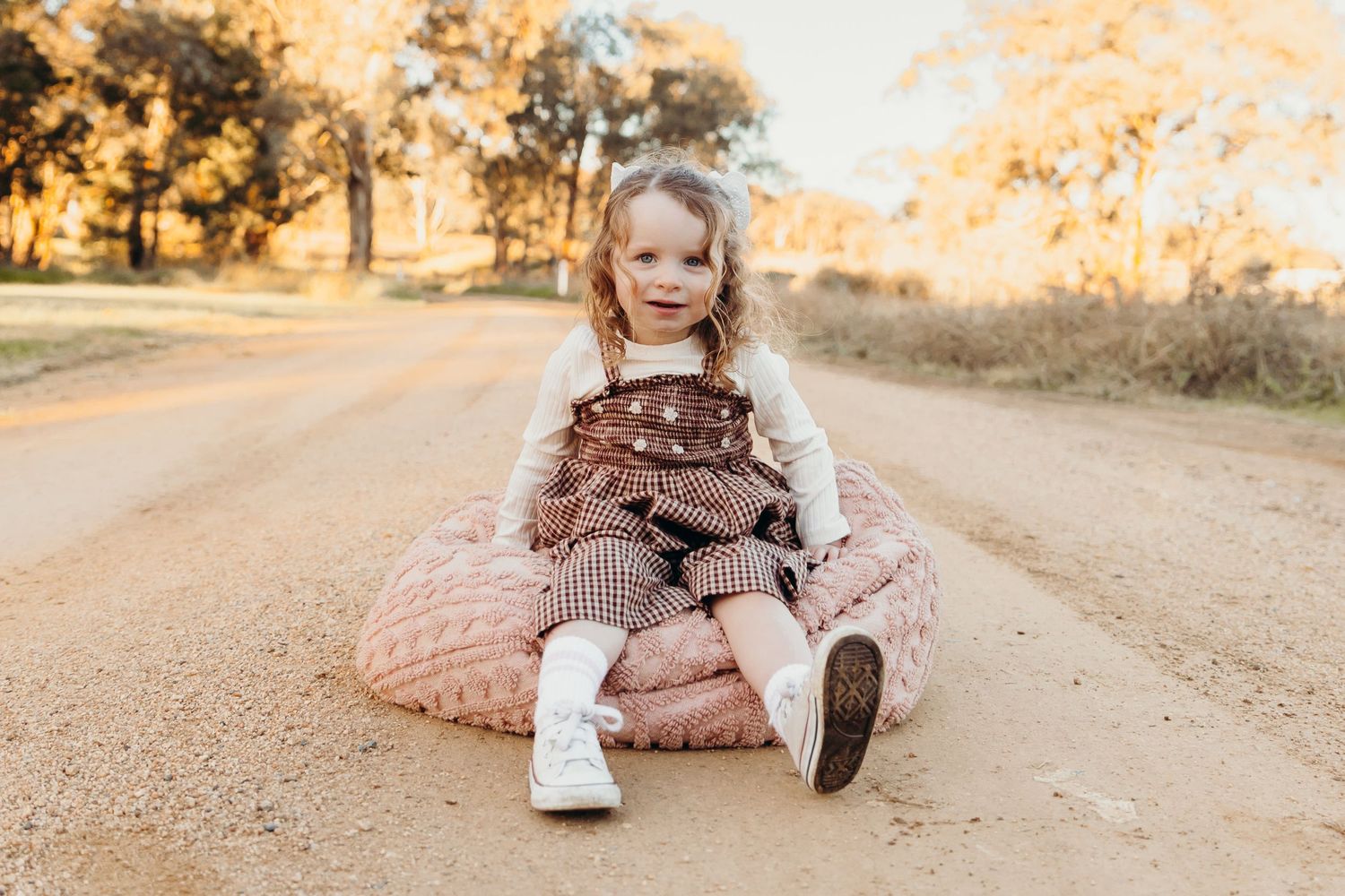 A young girl in a brown pinafore sits on a pink cushion on a dirt road surrounded by golden autumn trees.