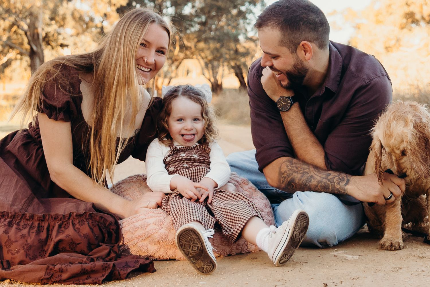 Parents and toddler laughing with their dog on a dirt path.