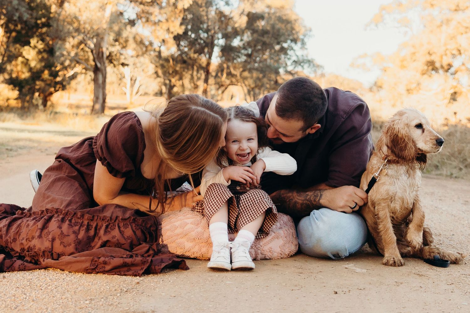Parents kissing smiling toddler while dog sits beside them.