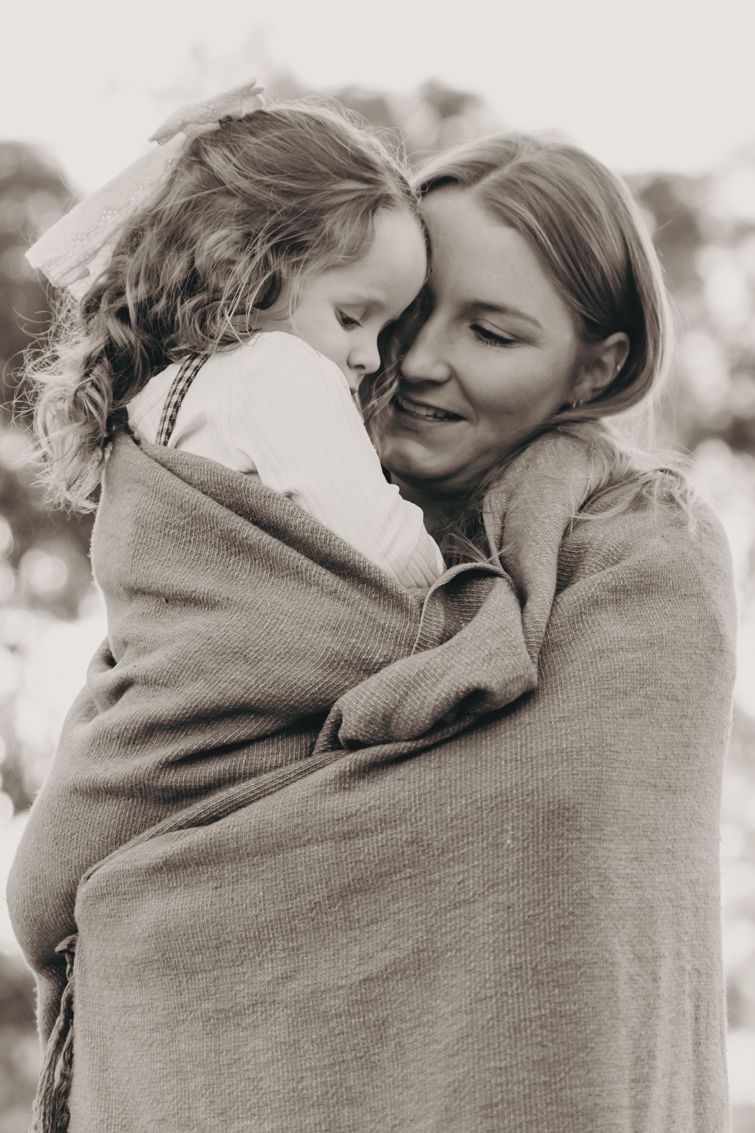 A tender black and white photo of a smiling mother wrapped in a blanket cuddling her young daughter outdoors.
