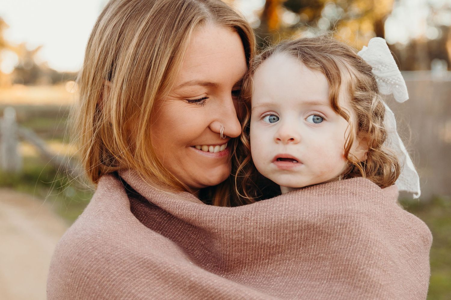 A smiling mother wrapped in a pink blanket cuddles her blue-eyed toddler girl outdoors.