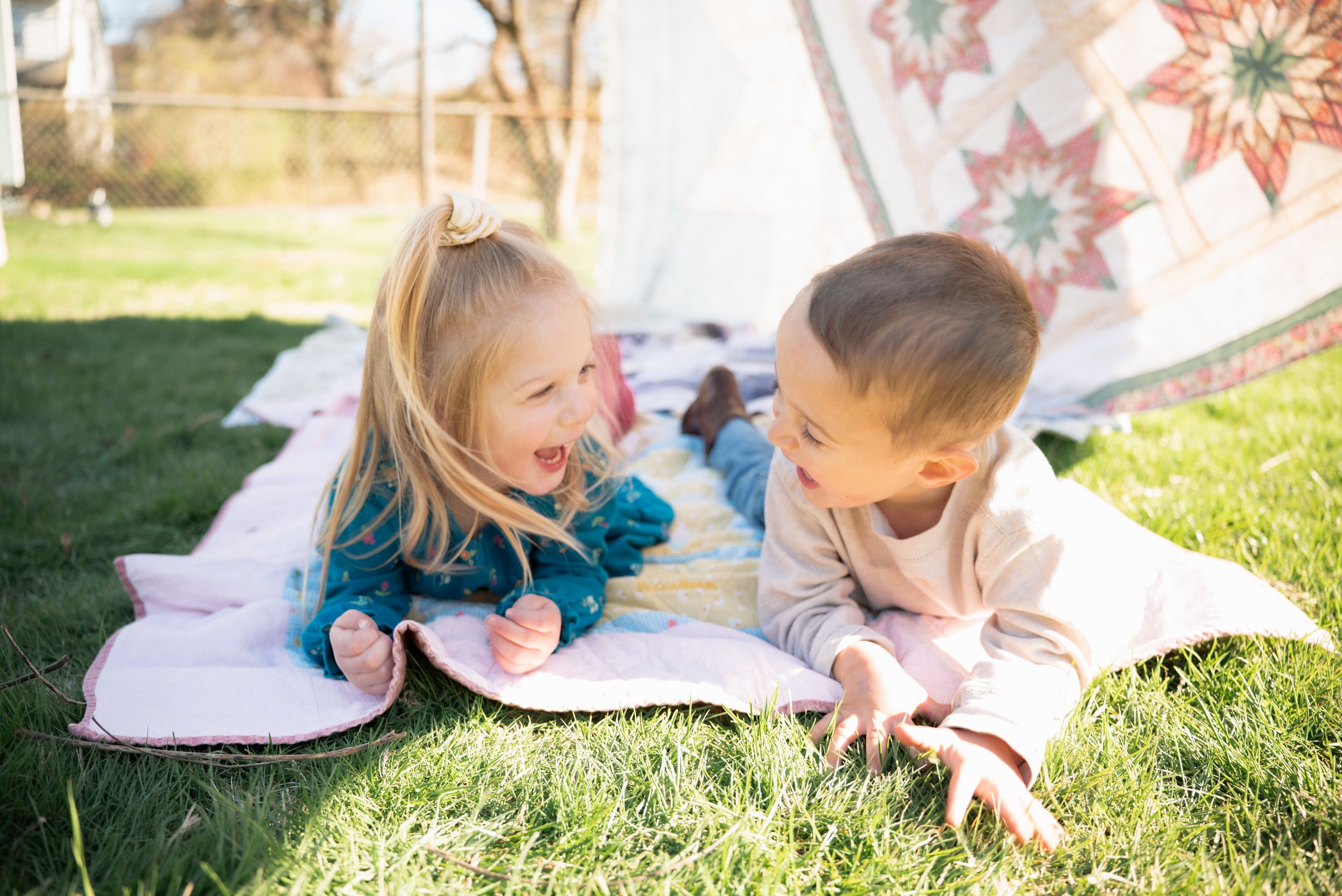 Two young children laughing and lying on a quilt blanket on green grass near a decorative tent outdoors.