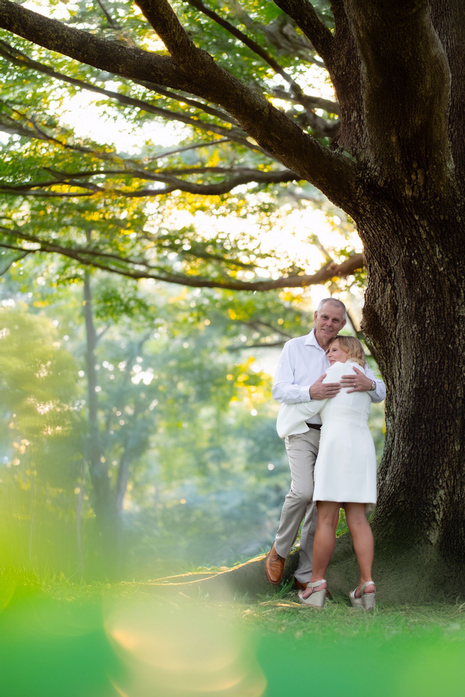 Couple in white outfits embracing under a large tree in a lush green park setting.