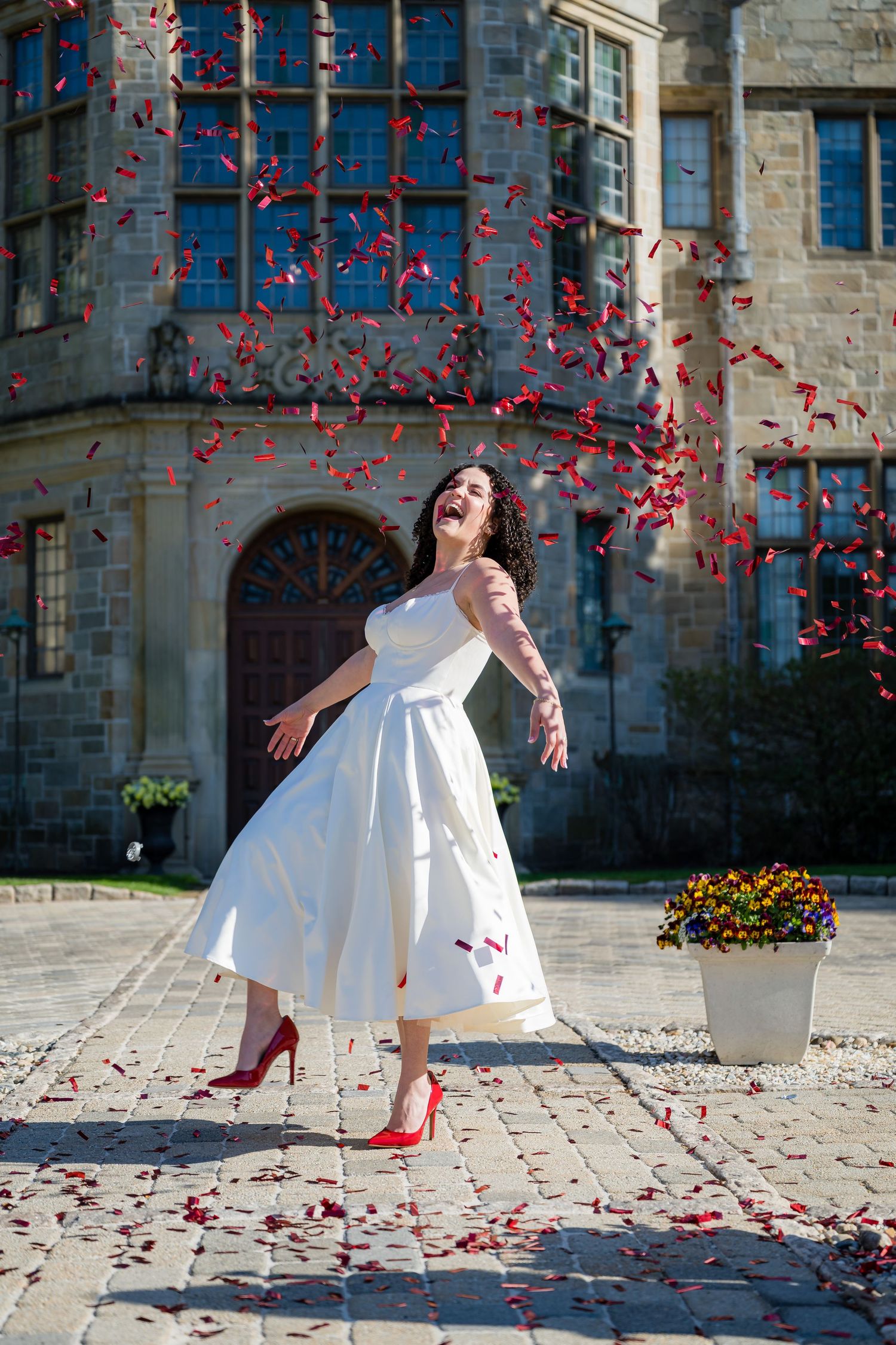Joyful bride in white midi dress and red heels twirling as red confetti falls outside a stone manor.