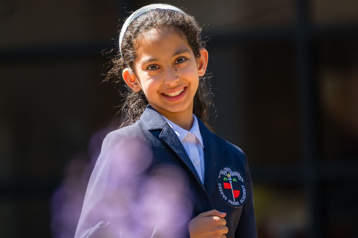 Smiling schoolgirl in navy blue uniform with emblem badge, wearing white headband, standing outdoors in bright sunlight.
