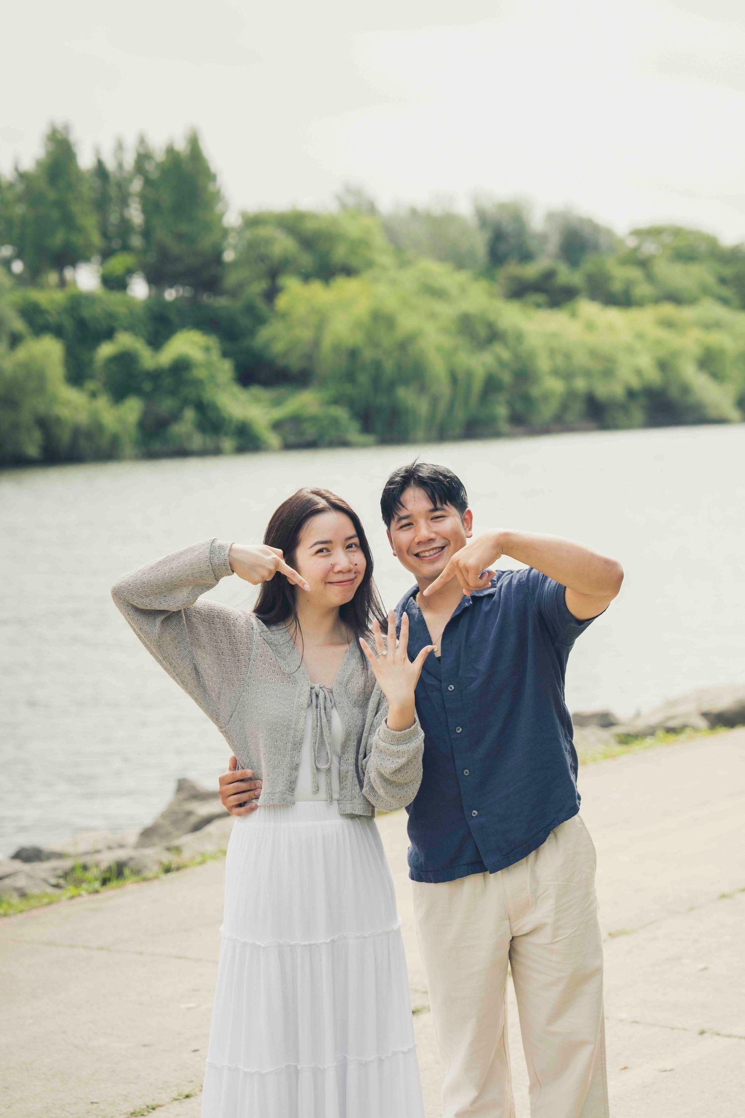 Happy Asian couple posing by a lake, showing off rings, surrounded by lush green trees in a park setting.
