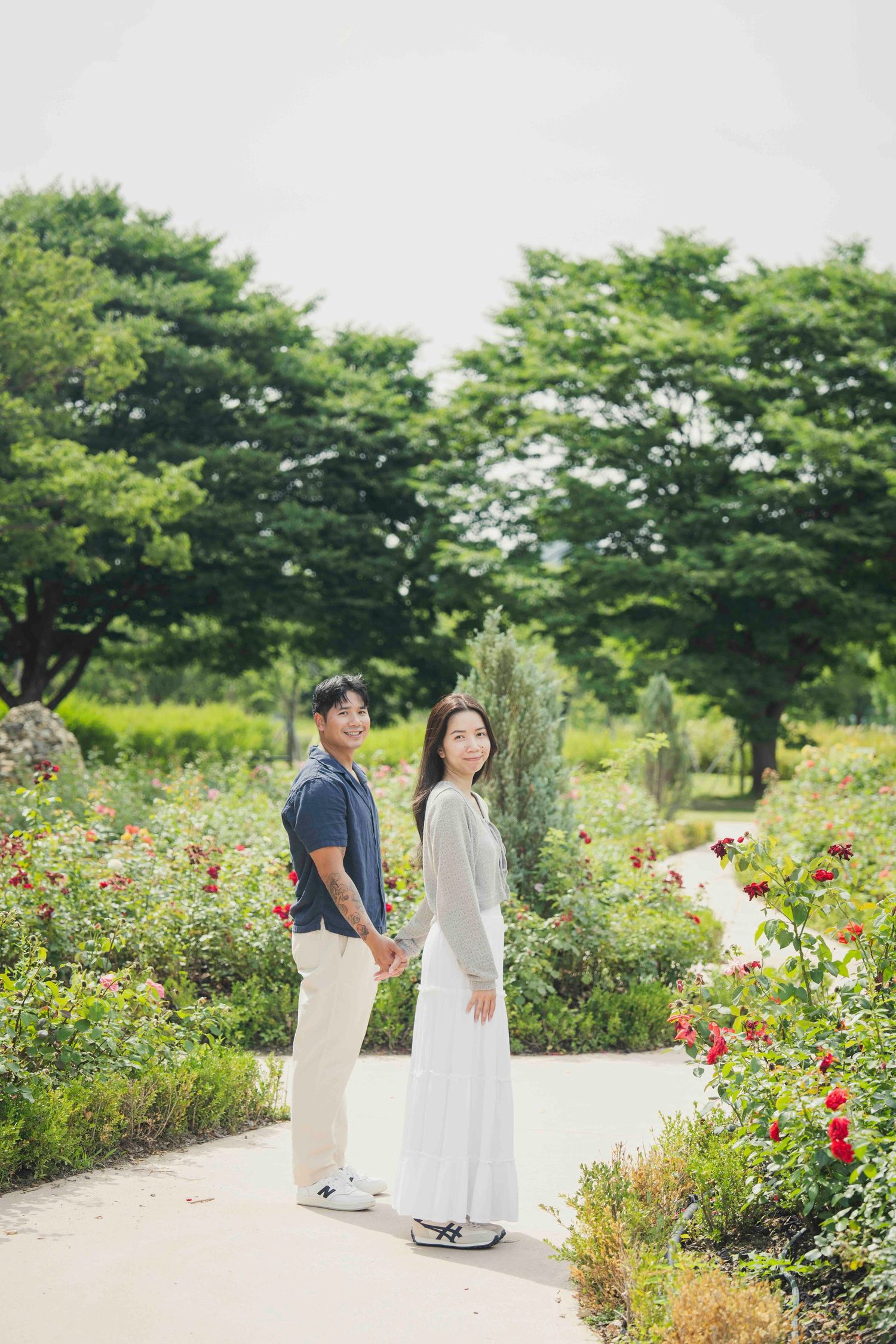 A couple holds hands on a garden path surrounded by colorful roses and lush green trees on a sunny day.