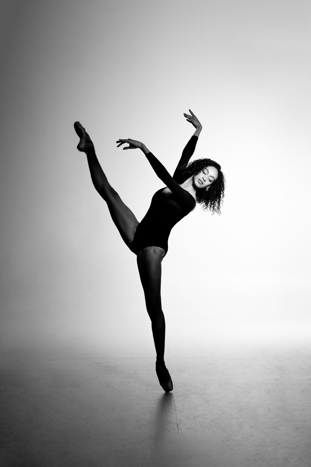 Black and white photo of a graceful ballet dancer balancing on one leg with arms raised in a studio.