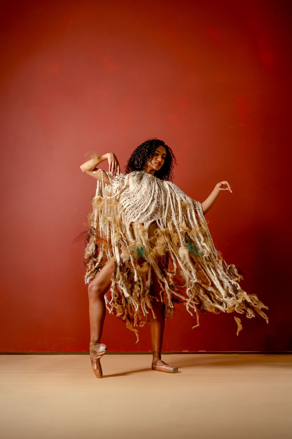 A dancer in a flowing fringed costume poses gracefully on pointe against a deep red background.