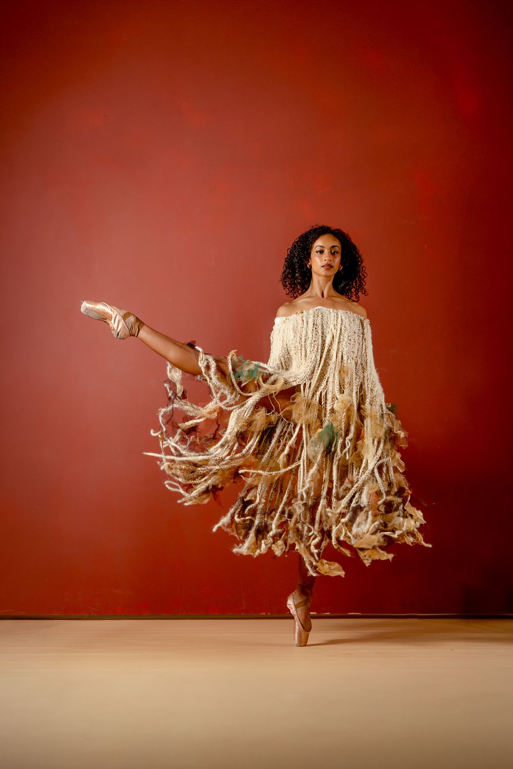 A ballerina in a flowing fringed dress poses en pointe against a red background.